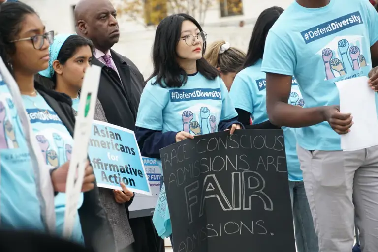 Color photo of students and advocates wearing blue shirts that say "#DefendDiversity" while standing outside the U.S. Supreme Court as the Court hears the cases SFFA v. Harvard and SFFA v. UNC on Oct. 31, 2022. The cases address race-conscious admissions policies. The students hold signs reading, "Race-conscious admissions are fair admissions" and "Asian Americans for affirmative action."