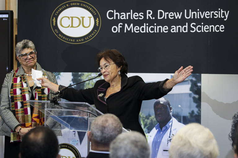 Color photo of Sylvia Drew Ivie speaking behind a podium, with her arms outstretched, at a press conference to announce the launch of the first historically Black four-year medical degree program at Charles R. Drew University of Medicine and Science on October 18, 2022. 