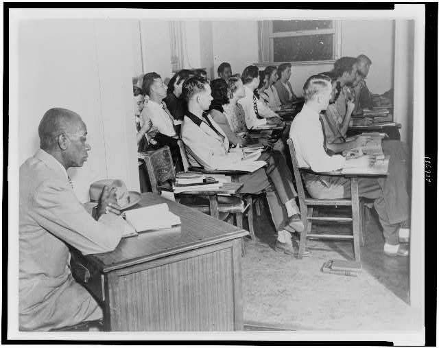 Black-and-white photo of George W. McLaurin, a 54-year-old Black man, sitting in an anteroom, apart from a classroom of white students, as he attends class at the University of Oklahoma in 1948. The university insisted that segregation be maintained, but a Supreme Court ruling forced the institution to accept McLaurin as a student.