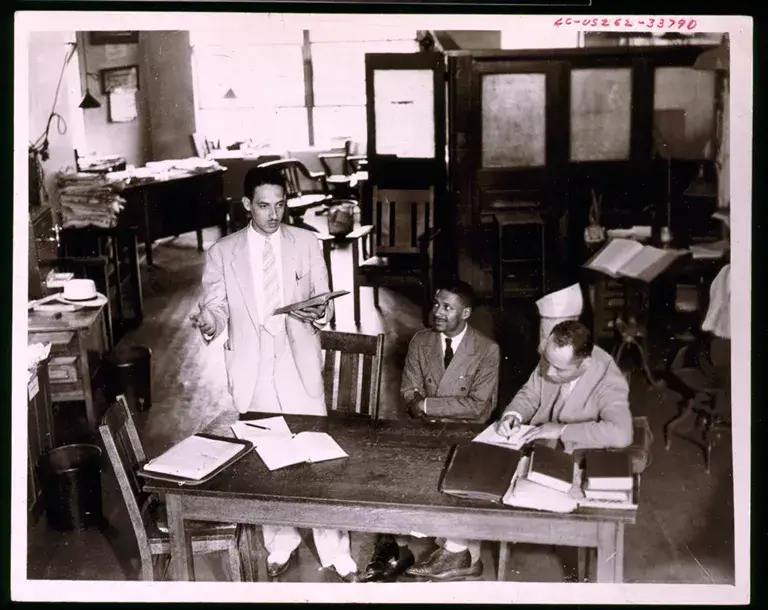 Black-and-white photo of a young Thurgood Marshall (left), standing in a meeting with two seated coworkers in an office.