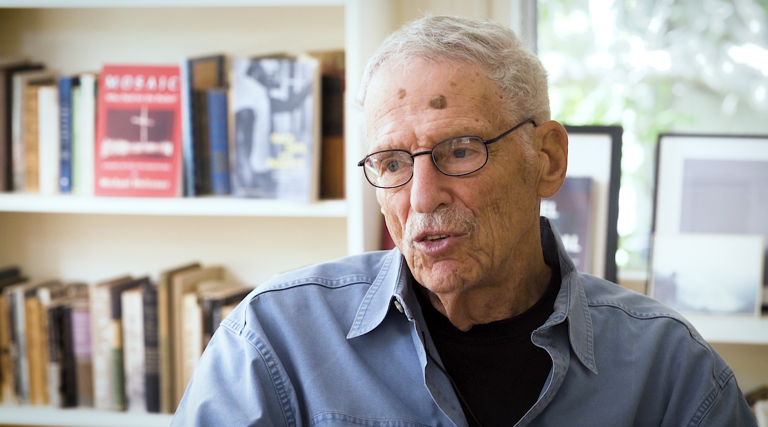 Color photo of Michael Meltsner, wearing glasses and a blue shirt, sitting in front of a bookshelf and a window during his oral history interview.