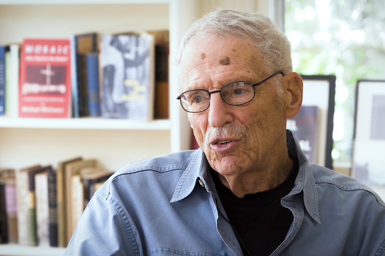 Color photo of Michael Meltsner, wearing glasses and a blue shirt, sitting in front of a bookshelf and a window during his oral history interview.