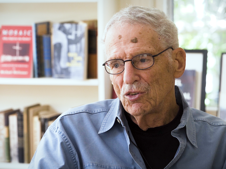 Color photo of Michael Meltsner, wearing glasses and a blue shirt, sitting in front of a bookshelf and a window during his oral history interview.