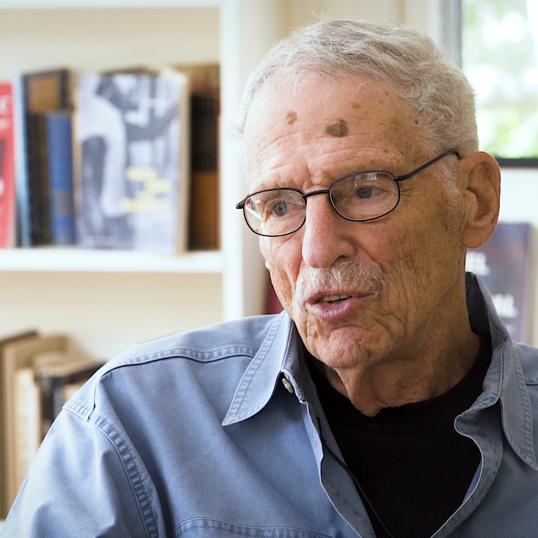 Color photo of Michael Meltsner, wearing glasses and a blue shirt, sitting in front of a bookshelf and a window during his oral history interview.