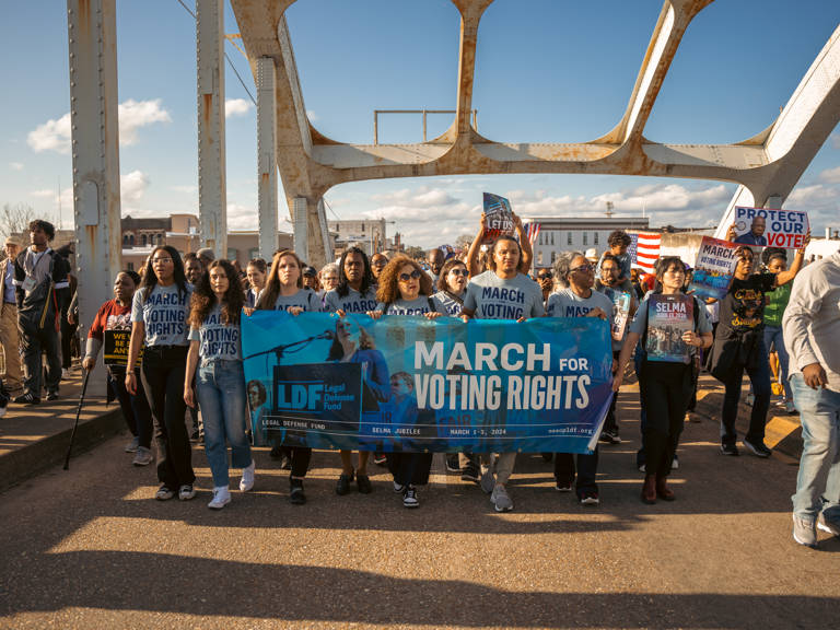 Color photo of a large group of LDF staff and other people marching across the Edmund Pettus Bridge while holding various voting rights signs and a banner reading, "LDF March for Voting Rights."