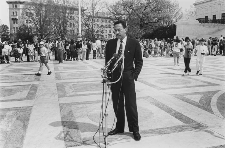 Black-and-white photo of Representative Mel Watt in a black suit standing outside in front of microphones. In the background are people walking. 