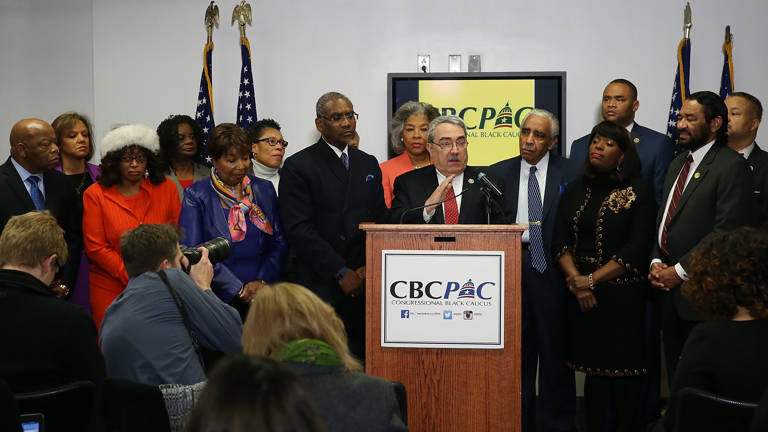 Color photo of U.S. Rep. G.K. Butterfield wearing a suit and speaking behind a podium while flanked by members of the Congressional Black Caucus during a news conference.