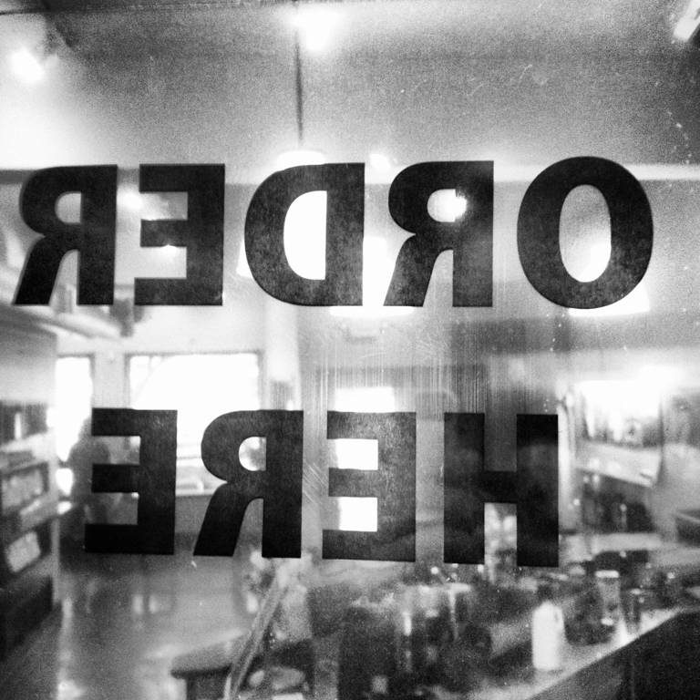 Black-and-white photo of a sign reading "Order Here" on a restaurant window.