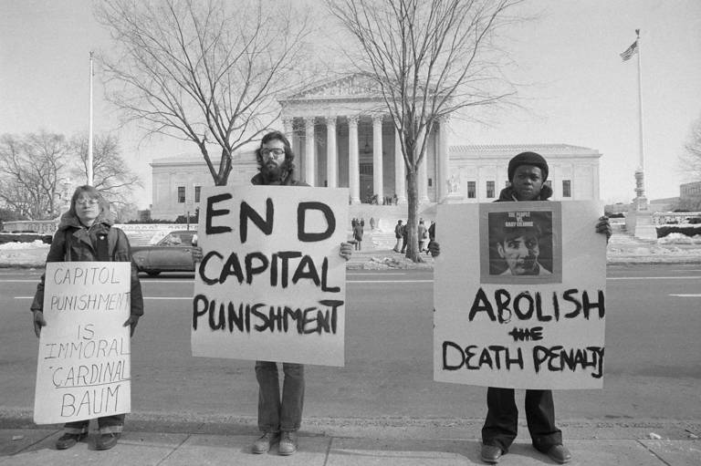 Black-and-white photo of three anti-death penalty demonstrators expressing their views with large signs reading, "Capital Punishment is Immoral Cardinal Baum," "End Capital Punishment," and "Abolish the Death Penalty" as they stand in front of the U.S. Supreme Court.