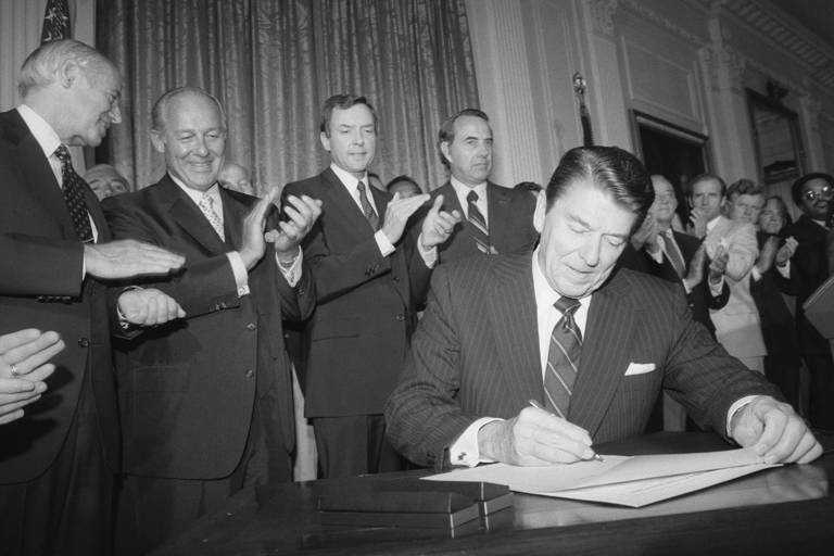 Black-and-white photo of President Ronald Reagan signing an extension of the Voting Rights Act while several men wearing suits stand behind him, clapping.