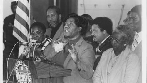 Black-and-white photo of Maggie Bozeman speaking behind a podium, flanked by several other people.