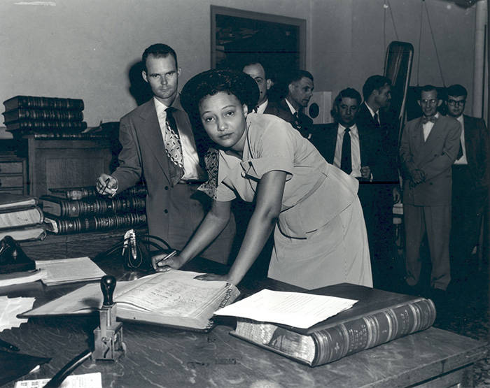 Black-and-white photo of American civil rights activist Ada Lois Sipuel Fisher signing the Register of Attorneys in Oklahoma City, Oklahoma, on July 1, 1952, as a line of men stand in the background.