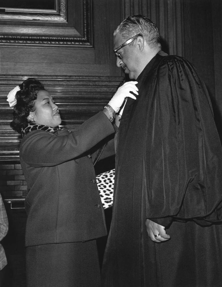 Black-and-white photo of Thurgood Marshall getting a last-minute check from his wife, American civil rights activist and historian Cecilia Suyat, before his swearing-in at the Supreme Court in 1967. Marshall is wearing a black judicial robe, and Suyat is helping Marshall adjust his robe.