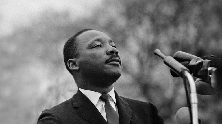 Black-and-white photo of the Rev. Martin Luther King Jr. speaking into microphones and wearing a suit.