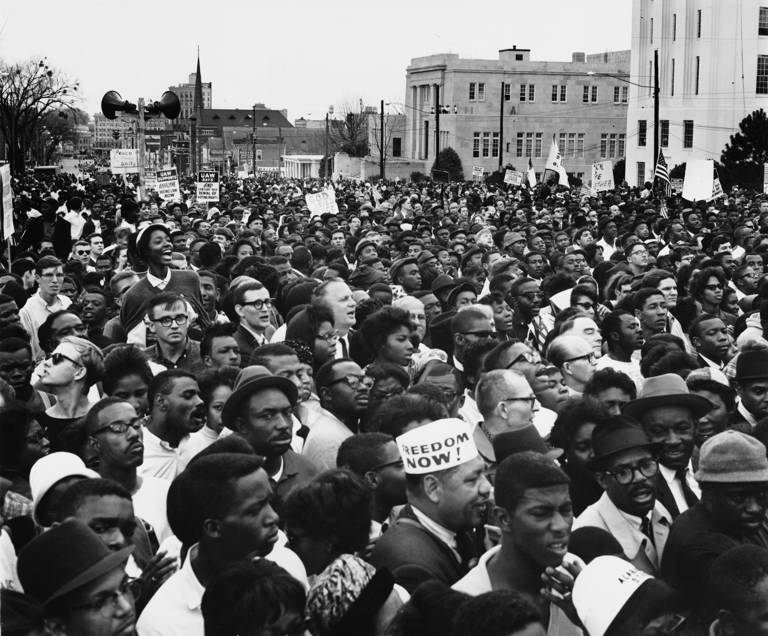 Black-and-white photo of a large crowd of people rallying in the street for voting rights, some holding signs. 