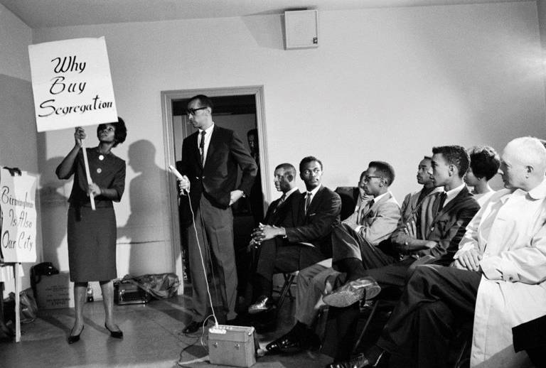 Black-and-white photo of Black pastor and civil rights leader Wyatt Tee Walker holding a microphone and speaking to civil rights activists gathered to discuss demonstration strategies against segregation in Birmingham, Alabama. On the left, a woman holds a placard reading, ‘Why Buy Segregation,’ during the Birmingham Campaign of 1963. Another placard reads: ‘Birmingham Is Also Our City.’