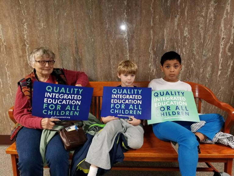 Color photo of three people, one adult and two children, sitting on a bench while holding signs reading, "Quality Integrated Education for All Children."