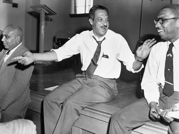 Black-and-white photo of Thurgood Marshall sitting at the edge of a stage and laughing with another man.